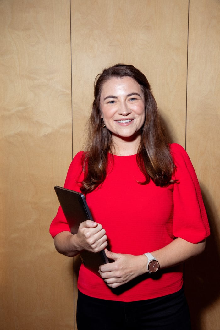 Eine Frau mit langen braunen Haaren lächelt breit in die Kamera. Sie trägt ein leuchtend rotes Oberteil und hält einen dunklen Ordner oder ein Tablet in der Hand. Sie steht vor einer hellen Holzwand oder -tafel.